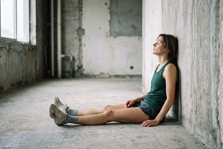 A runner sitting on a concrete floor after a session, legs extended, head tipped back against the wall, morning light across the room, completely still