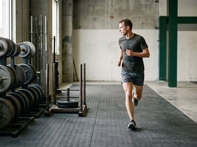 A lean distance runner mid-stride on an indoor floor, passing a rack of loaded barbells and a sled