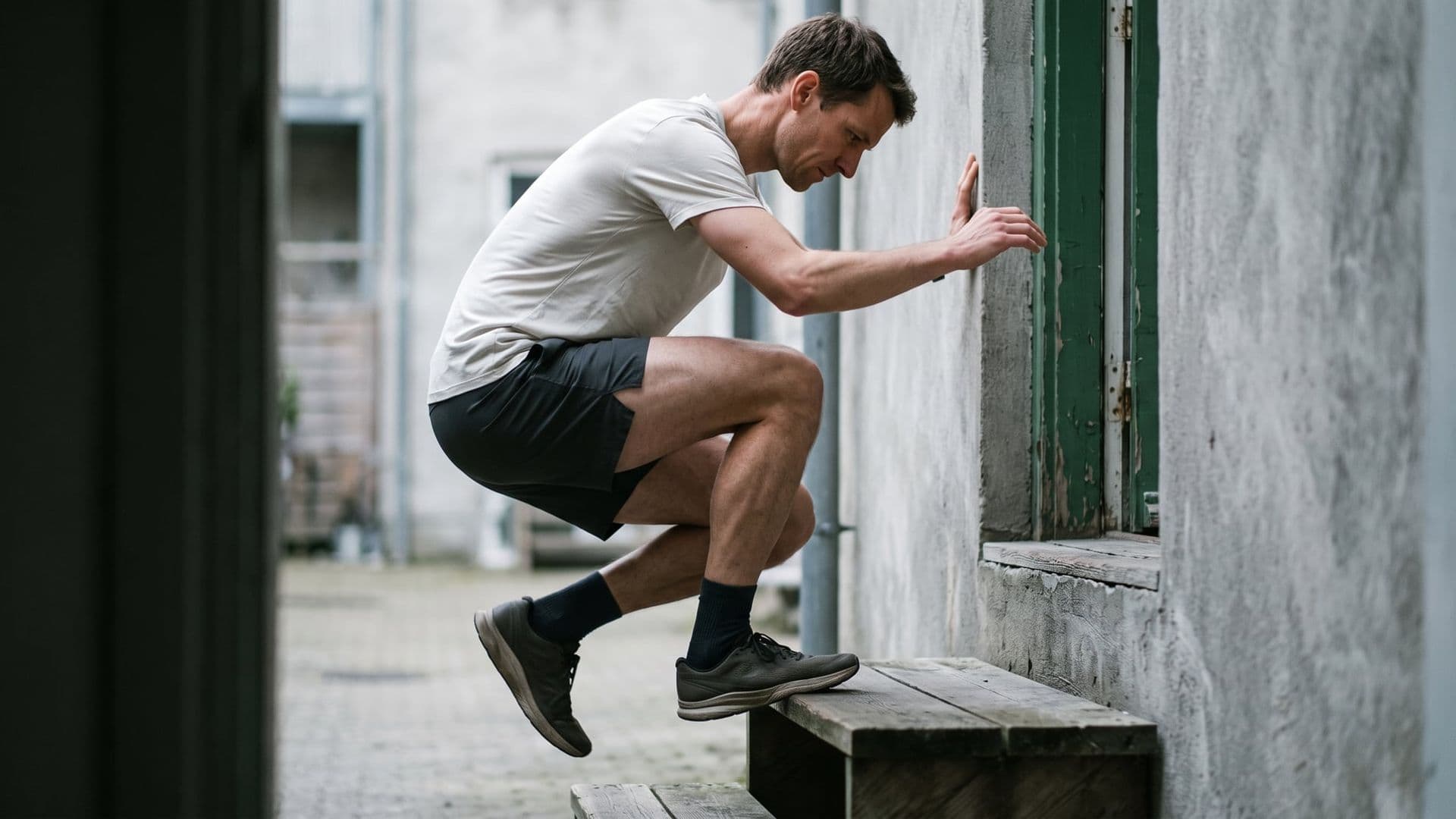 A close low-angle view of a runner performing a single-leg calf raise on the edge of a wooden step, one heel dropped below the step, hand resting lightly on a concrete wall for balance