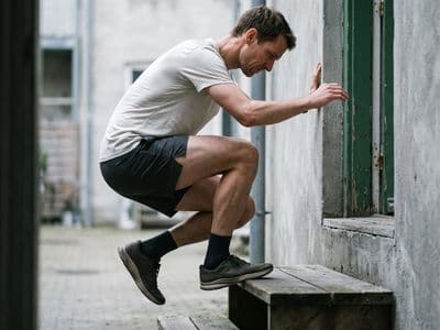 A close low-angle view of a runner performing a single-leg calf raise on the edge of a wooden step, one heel dropped below the step, hand resting lightly on a concrete wall for balance