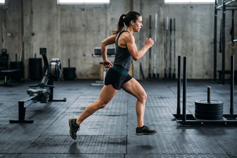 An athlete mid-stride running between stations on a rubber-matted indoor floor, an unbranded loaded sled and a rowing machine out of focus behind them